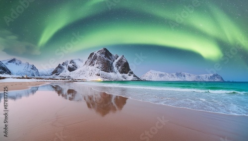 Aurora borealis over the sea, snowy mountains and city lights at night. Northern lights in Lofoten islands, Norway. Starry sky with polar lights. Winter landscape with aurora, reflection, sandy beach.