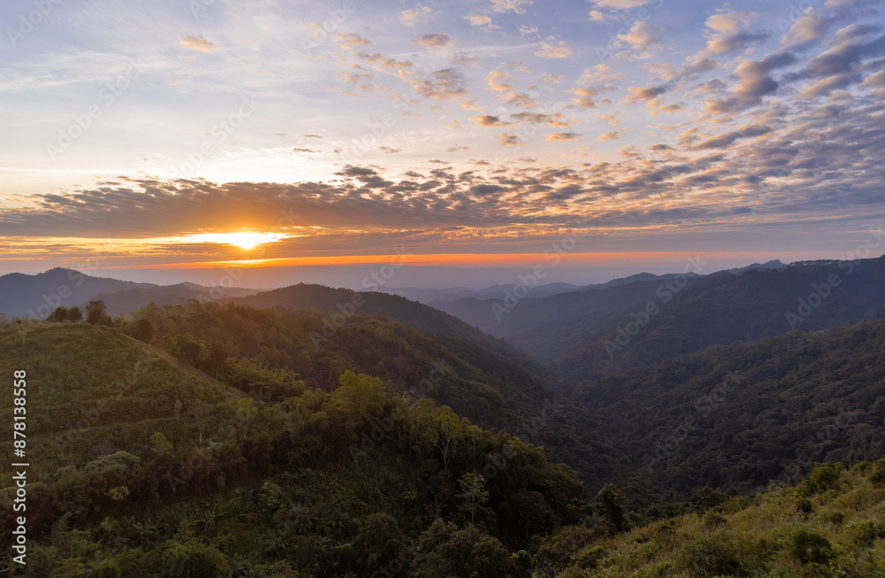 Beautiful landscape of a sunset or sunrise over a mountainous area. The sky is partially cloudy, and the scene shows a vast expanse of hills covered with trees and vegetation.