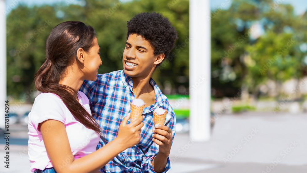 Fototapeta premium A teen black couple stand close together and smile as they each hold a waffle cone ice cream. They are standing outside on a sunny day, copy space