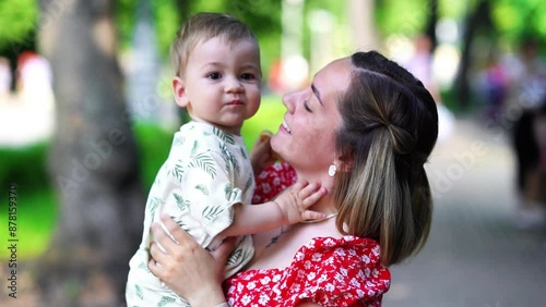 Cheerful young mother with her toddler son in a summer park, having fun together. They are smiling, playing, and sharing affectionate moments, with the mother lifting and kissing her son on the cheek
