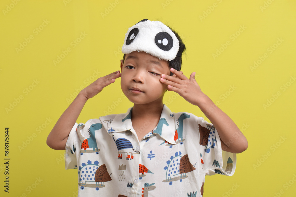 Portrait of Asian child wearing pajamas and sleep mask rubbing his eyes isolated on yellow background. Child wants to sleep. Healthy sleep concept. Copy space Portrait of Asian child wearing pajamas and sleep mask rubbing his eyes isolated on yellow background. Child wants to sleep. Healthy sleep concept. Copy space