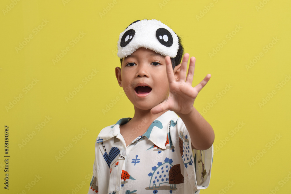 Portrait of Asian child wearing pajamas and sleeping mask isolated on yellow background. Child wants to sleep. Healthy sleep concept. Copy space