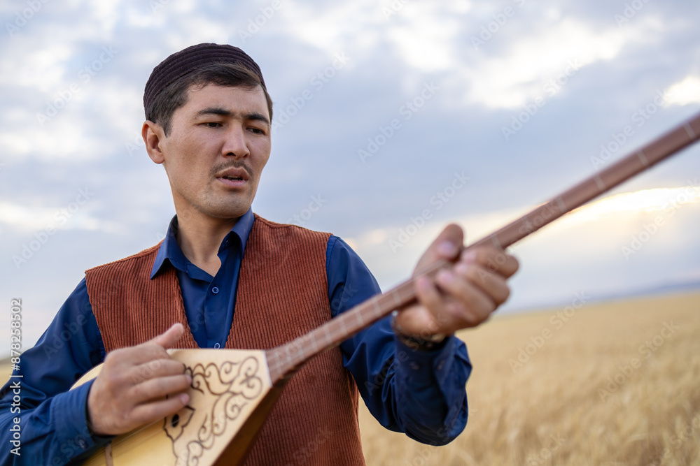 Fototapeta premium Kazakh, a man in Kazakh national dress playing dombra in the steppe. Kazakhstan. Central Asia. Nomads. | Казах, мужчина в казахской национальной одежде играет на домбре в степи. Казахстан.Средняя Азия