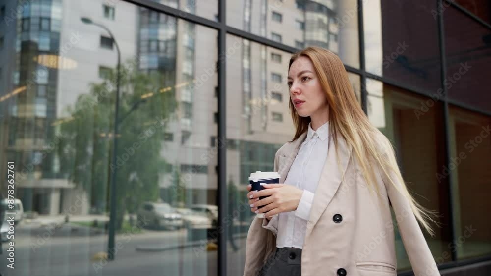 A young businesswoman walking with a coffee cup in hand, displaying a focused and determined expression. She is dressed in a beige coat and white shirt, perfect for a professional environment. A young businesswoman walking with a coffee cup in hand, displaying a focused and determined expression. She is dressed in a beige coat and white shirt, perfect for a professional environment.