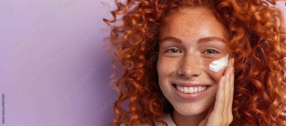 Fototapeta premium Portrait of a beautiful young woman with red curly hair smiling and applying cream to her face against a purple background, shown in closeup