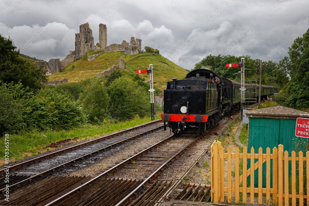Fototapeta premium A steam train pulling into Corfe Castle Dorset UK with storm clouds in the sky and Corfe Castle ruins in the background