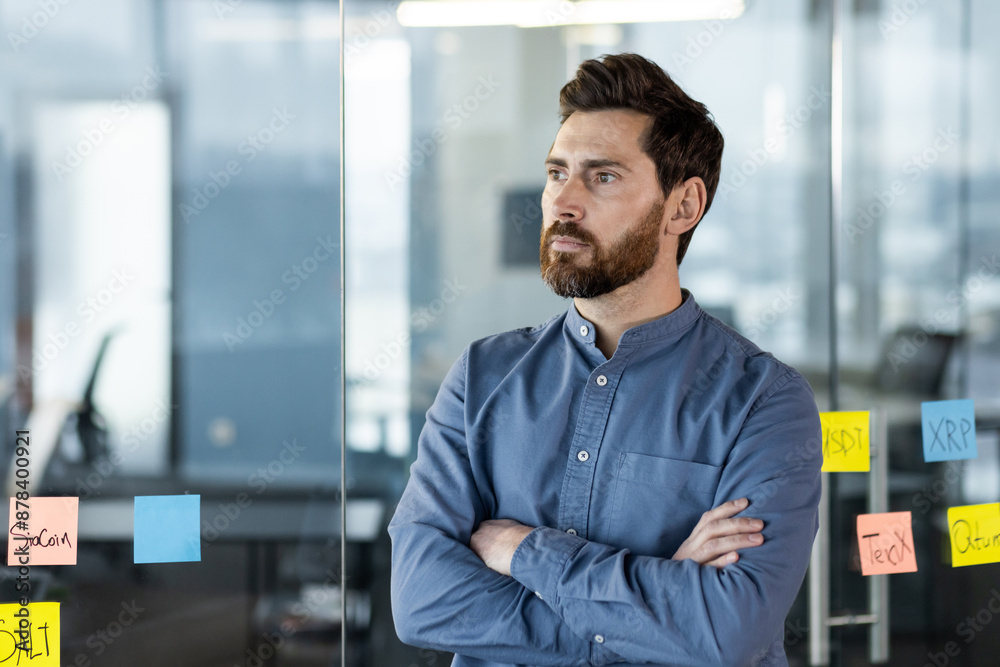 A young businessman man stands in a modern office in a blue shirt, crosses his arms on his chest, looks seriously and thoughtfully to the side A young businessman man stands in a modern office in a blue shirt, crosses his arms on his chest, looks seriously and thoughtfully to the side