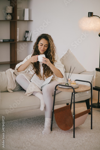 Young woman reading book and drinking tea, sitting on sofa in a cozy living room.