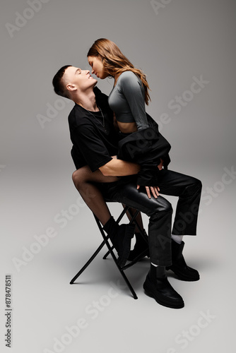 A man and a woman, a young couple in love, sit together on a chair in a studio with a grey background.