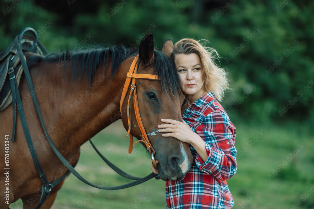 Obraz premium Happy blonde with horse in forest. Woman and a horse walking through the field during the day. Dressed in a plaid shirt and black leggings.