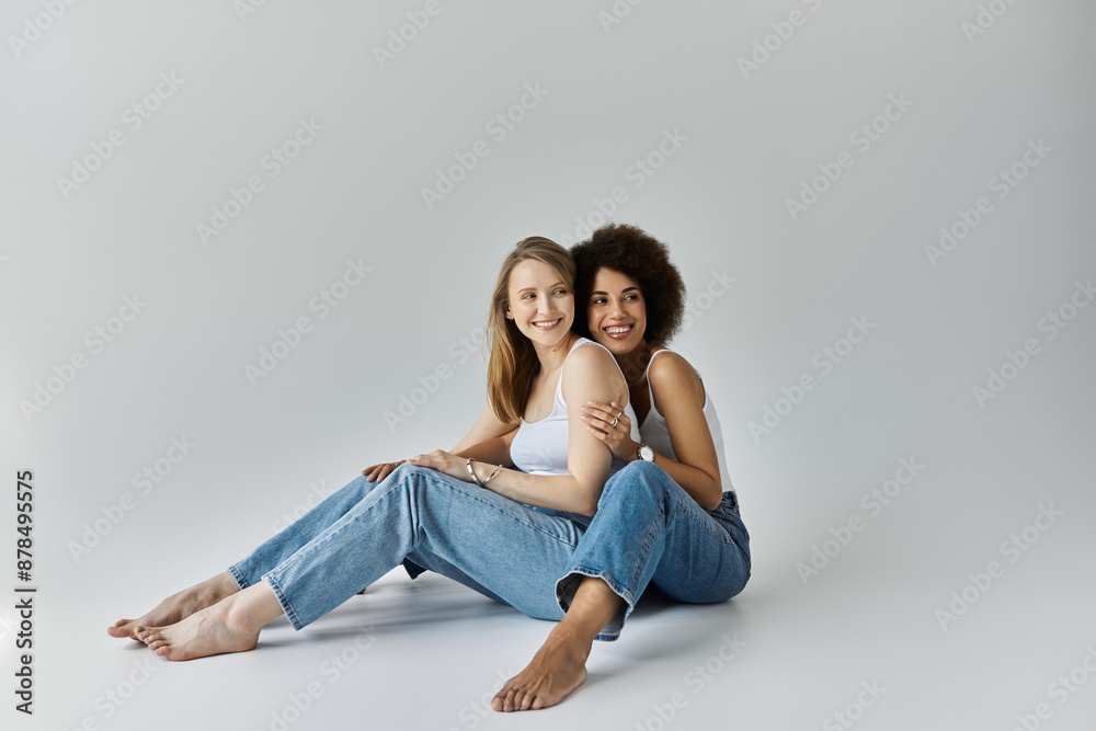 Two women, one with dark skin and one with fair skin, sitting together with arms around each other, gazing affectionately Two women, one with dark skin and one with fair skin, sitting together with arms around each other, gazing affectionately