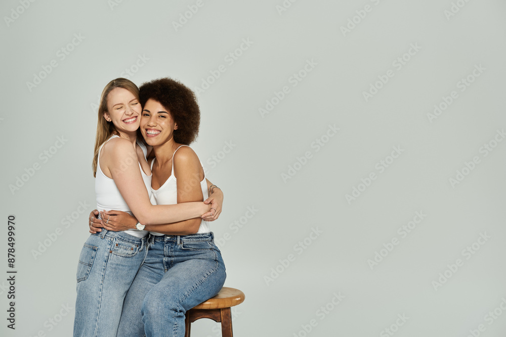 A lesbian couple in white tank tops and jeans hug and smile in a studio setting. A lesbian couple in white tank tops and jeans hug and smile in a studio setting.