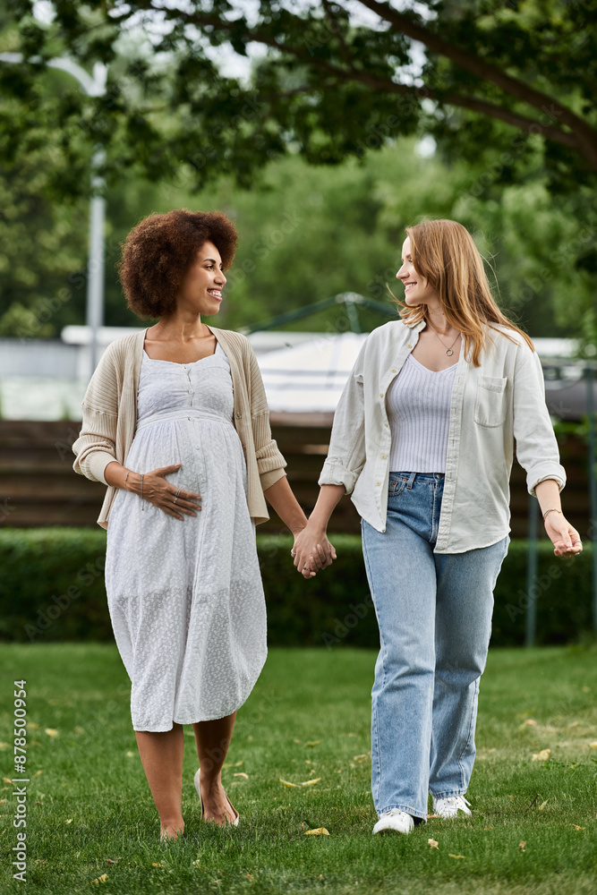 A pregnant Black woman and her partner walk hand-in-hand in a park, smiling and looking at each other. A pregnant Black woman and her partner walk hand-in-hand in a park, smiling and looking at each other.
