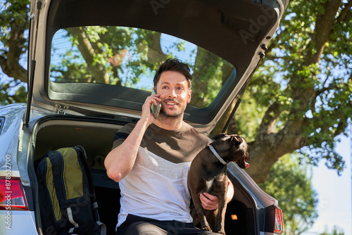 man sitting in the trunk talking on the phone. he has a dark dog in his arms. photo