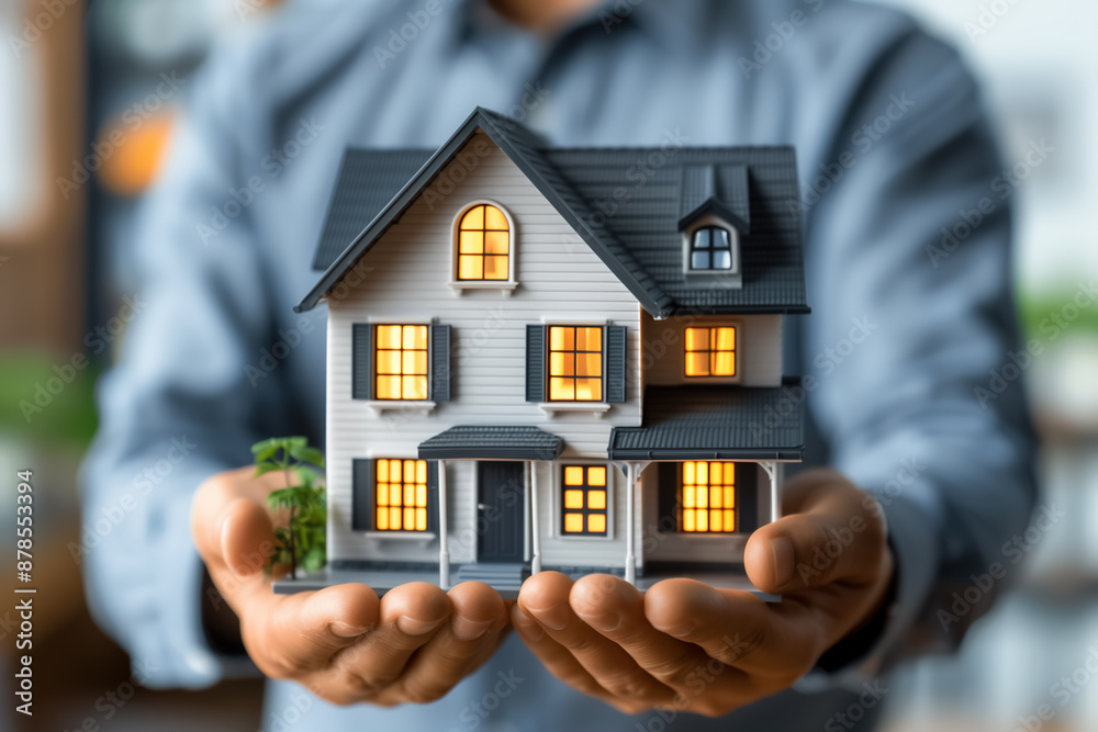 A man holds a detailed realistic model of a two-story house with a sloping roof. A man holds a detailed realistic model of a two-story house with a sloping roof.