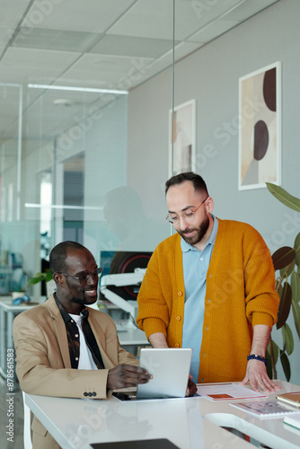 Two diverse male colleagues, Caucasian man and Black man, smiling while discussing project details on digital tablet in modern office