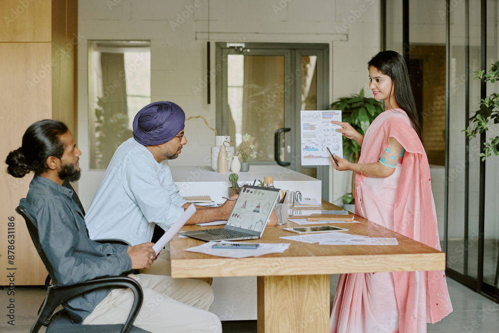 Female Indian teamlead demonstrating future plans and goals to male subordinates while they sitting at wooden work desk Female Indian teamlead demonstrating future plans and goals to male subordinates while they sitting at wooden work desk