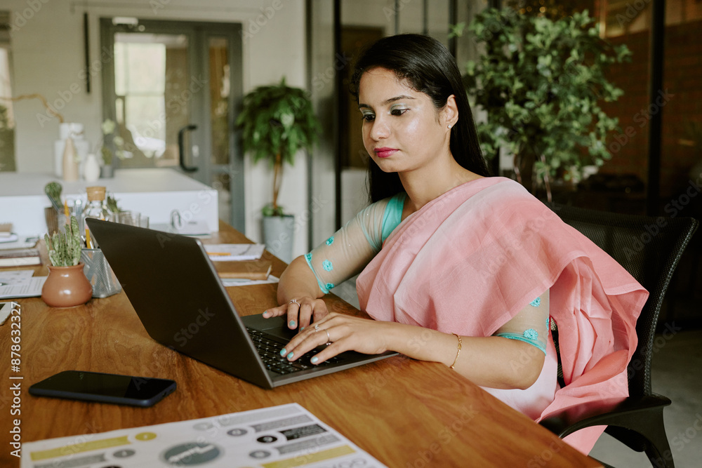 Young beautiful female IT specialist wearing traditional Indian dress sitting at wooden table and searching information in Internet while in office Young beautiful female IT specialist wearing traditional Indian dress sitting at wooden table and searching information in Internet while in office