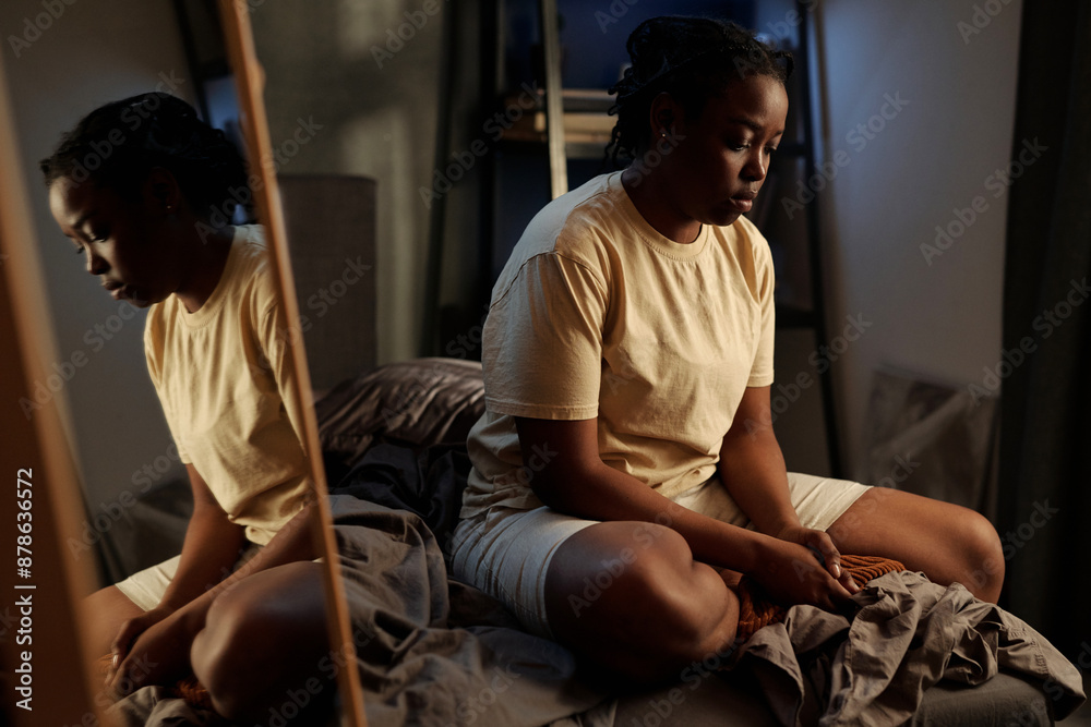 Young unhappy African American woman sitting on bed by mirror and suffering from insomnia caused by post traumatic syndrome Young unhappy African American woman sitting on bed by mirror and suffering from insomnia caused by post traumatic syndrome
