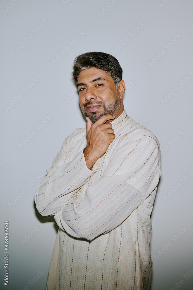 Portrait of young Indian man dressed in patterned creamy shirt holding his chin and smirking with squint while sitting for photo Portrait of young Indian man dressed in patterned creamy shirt holding his chin and smirking with squint while sitting for photo