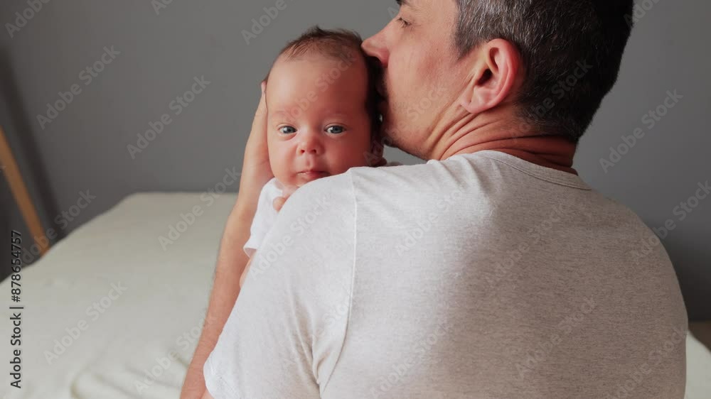 Anonymous man dressed in white T-shirt posing in bedroom interior rocking his newborn baby in hands kissing tiny child hugging his cute son or daughter calming down infant kid Anonymous man dressed in white T-shirt posing in bedroom interior rocking his newborn baby in hands kissing tiny child hugging his cute son or daughter calming down infant kid