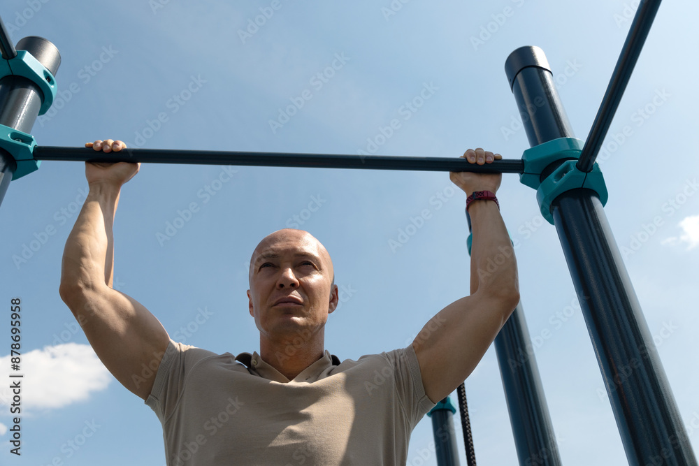 Naklejka premium Handsome man does pull-ups on horizontal bar at street on sports ground. Outdoors. Summer. Workout. Morning routine.