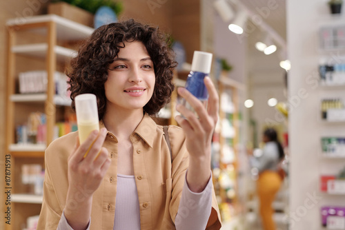 Happy young brunette woman in casualwear choosing makeup remover while standing in cosmetic supermarket and looking at bottle photo