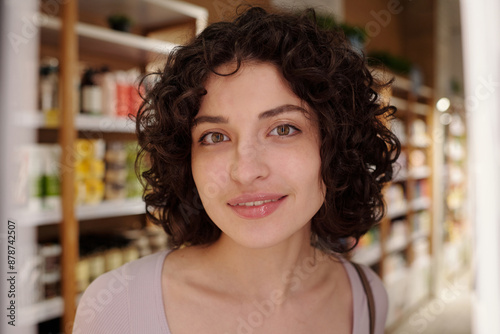 Young gorgeous brunette woman with curly hair looking at camera after applying new balm on lips while standing in cosmetic shop