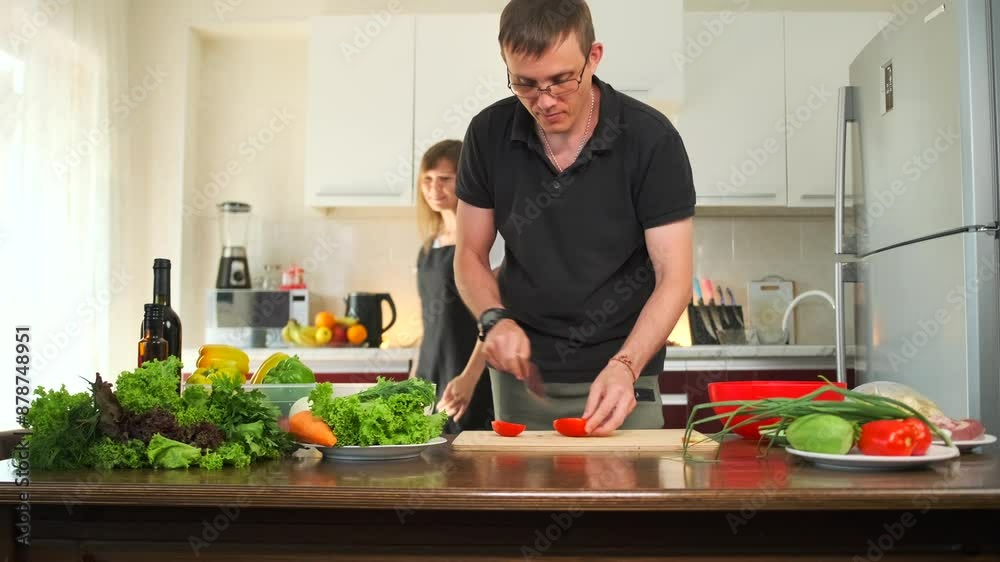 Man cutting tomato with a sharp knife, next to his wife at the kitchen counter with lying fresh leafy vegetables bell peppers while preparing dinner together. Man cutting tomato with a sharp knife, next to his wife at the kitchen counter with lying fresh leafy vegetables bell peppers while preparing dinner together.