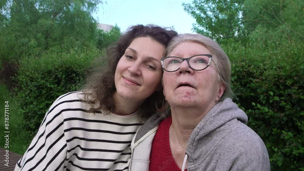 An adult daughter and an elderly mother with dementia on a walk in the park. The women are sitting on a bench and carefully holding each other's hands. An adult daughter and an elderly mother with dementia on a walk in the park. The women are sitting on a bench and carefully holding each other's hands.