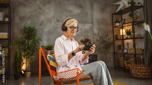 Mature woman sit on chair with headphones listen music on mobile phone