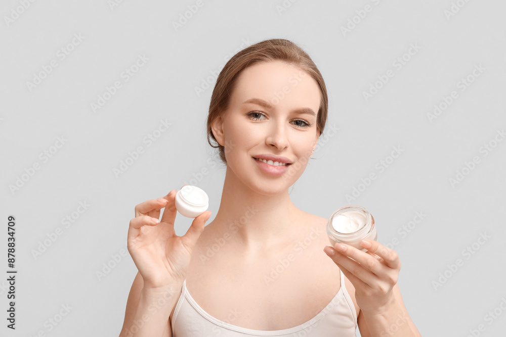 Beautiful young woman with jars of facial cream on grey background
