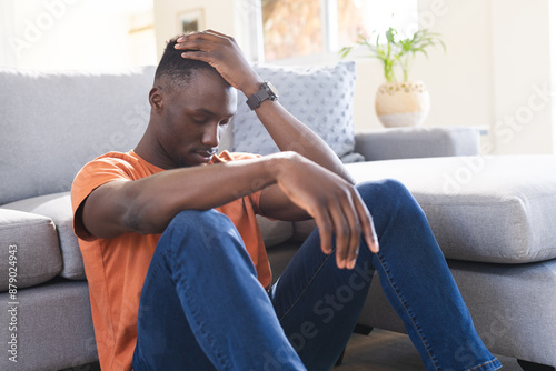 Thoughtful african american man sitting on floor holding head in sunny living room