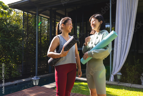 Happy asian female friends holding yoga mats and talking in garden at home photo