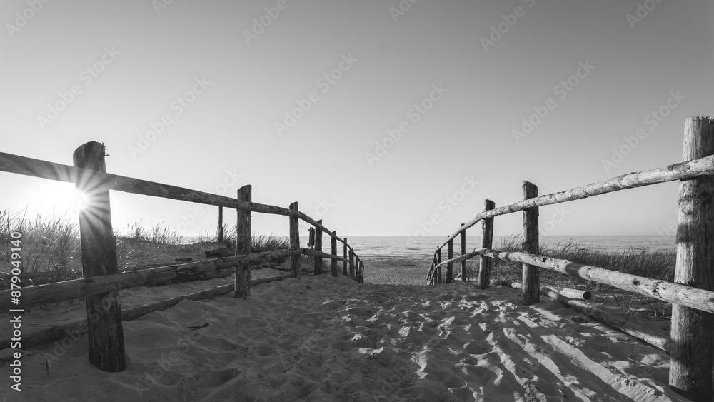 Black and white photo. Sandy entrance to the beach. Sunset. Wooden balustrades. Footprints in the sand. Dunes covered with grass. Slajszewo, Poland. Baltic Sea