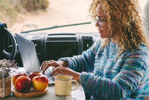 One modern female tourist working on laptop sitting inside a modern camper van with beach view parking in background. Woman using computer inside vehicle. Concept of vanlife and travel lifestyle