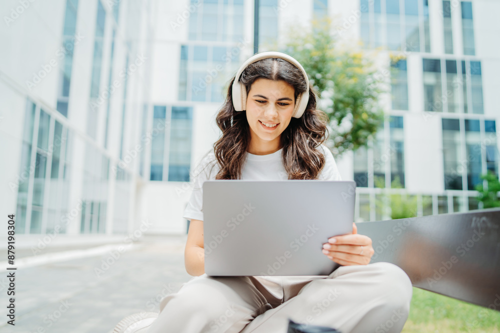 Fototapeta premium One beautiful young caucasian woman student sitting on bench near university listening to music and studying on laptop 