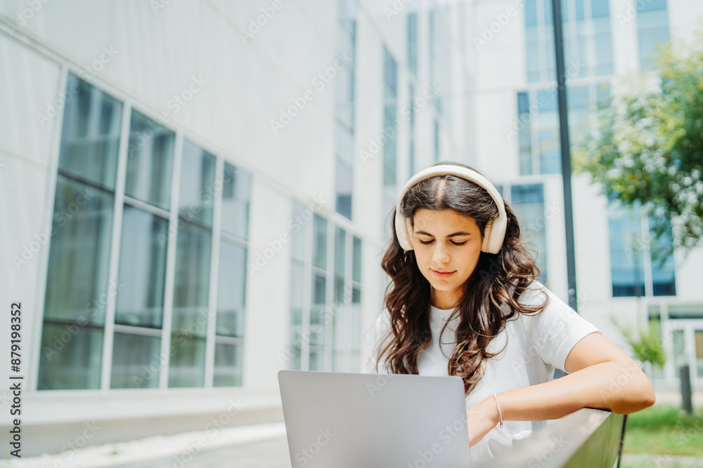 Fototapeta premium One beautiful young caucasian woman student sitting on bench near university listening to music and studying on laptop