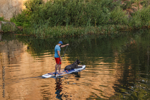 young man swimming standing on SUP board with dog of fighting breed, lake, river, dawn. The person resting with his pet gray Cane Corso. photo