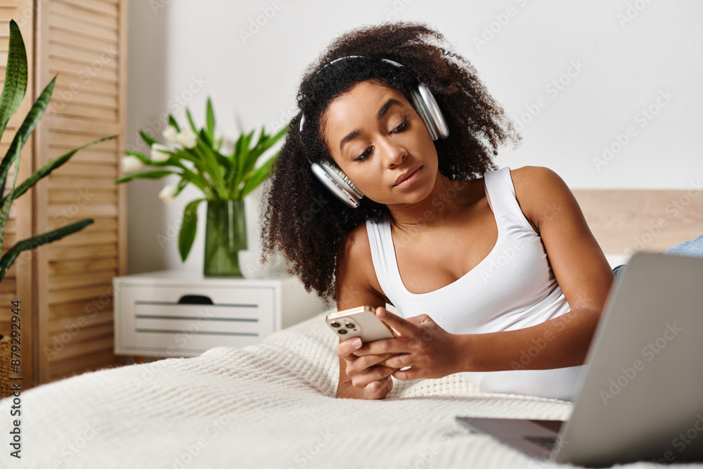 A curly African American woman in a tank top relaxes in bed, listening to music on her phone in a modern bedroom. A curly African American woman in a tank top relaxes in bed, listening to music on her phone in a modern bedroom.