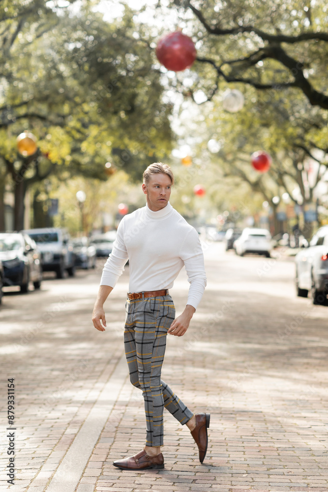 Fototapeta premium A blonde man in a white turtleneck and plaid pants poses on a street in Florida.