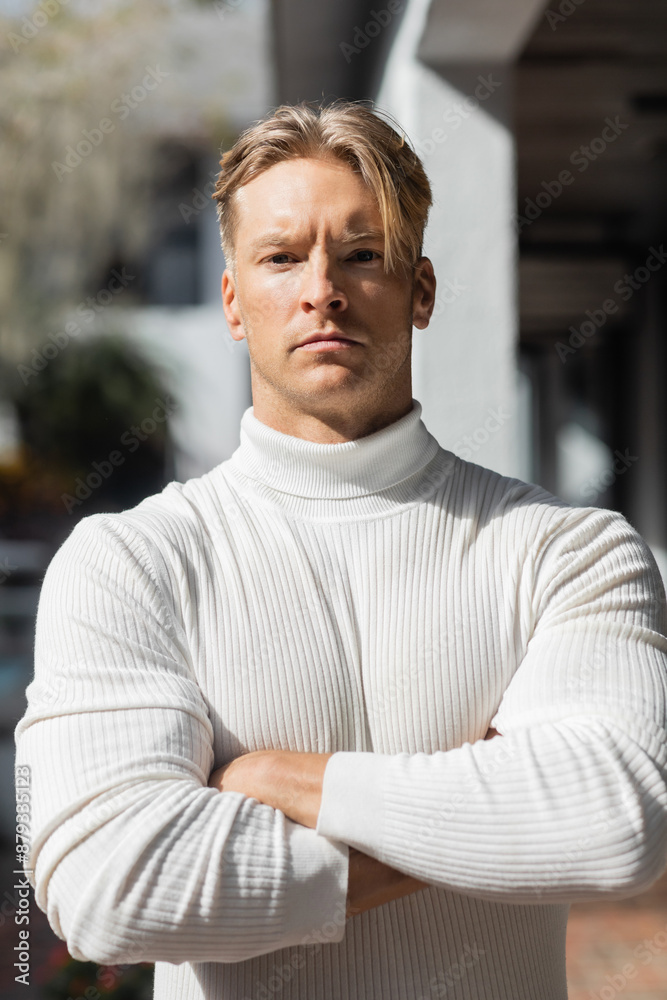 Fototapeta premium A handsome blonde man poses on a Florida street, wearing a white turtleneck sweater and looking directly at the camera.
