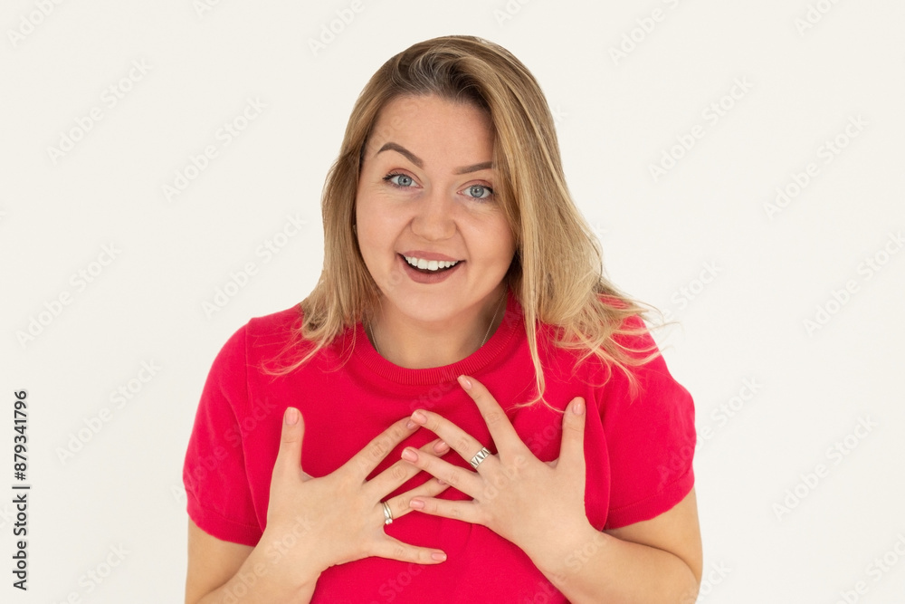 Caucasian young cheerful girl modestly laughs clasping her hands to chest on white background. Woman smiles broadly, wearing red t-shirt. Reaction to happiness, concept Caucasian young cheerful girl modestly laughs clasping her hands to chest on white background. Woman smiles broadly, wearing red t-shirt. Reaction to happiness, concept