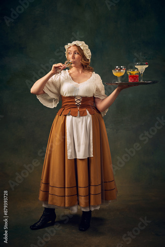 Portrait of woman in historical attire, drinking cocktail and holding a tray with various alcoholic drinks, set against vintage green background. Concept of history, medieval bar, restaurant