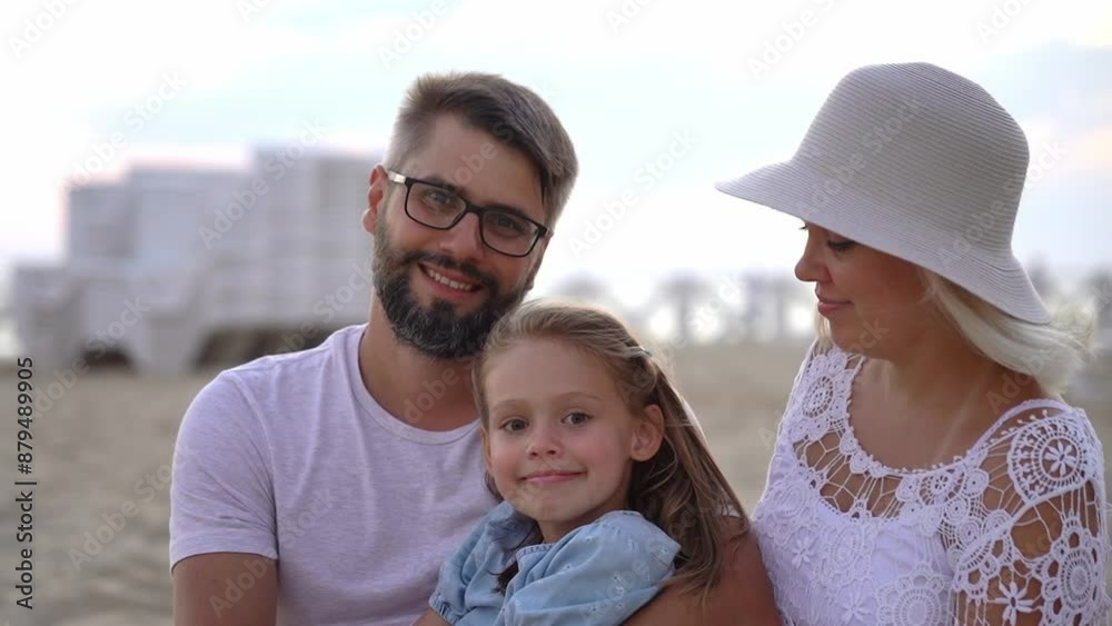 Portrait of happy family looks at camera and smiles on beach by sea at sunset. Father, mother and child in summer outdoors. Travel, vacation, holiday, tourism. Close up Portrait of happy family looks at camera and smiles on beach by sea at sunset. Father, mother and child in summer outdoors. Travel, vacation, holiday, tourism. Close up