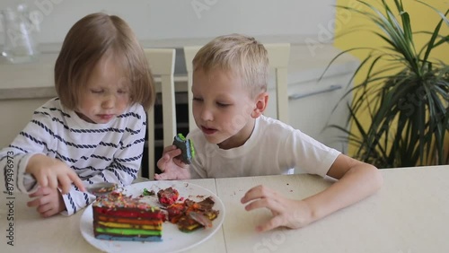 The older brother and little sister are laughing and eating a multicolored cake.
