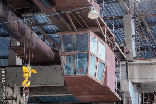 Overhead crane with a linear cross-beam and a yellow hook in the shop of the machine-building plant. Crane operator's cabin and trolley of the boom crane. Industrial production concept.