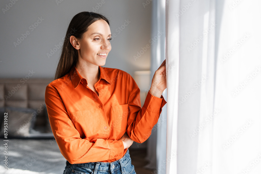 Smiling woman wearing orange shirt , looking at window while standing at home Smiling woman wearing orange shirt , looking at window while standing at home
