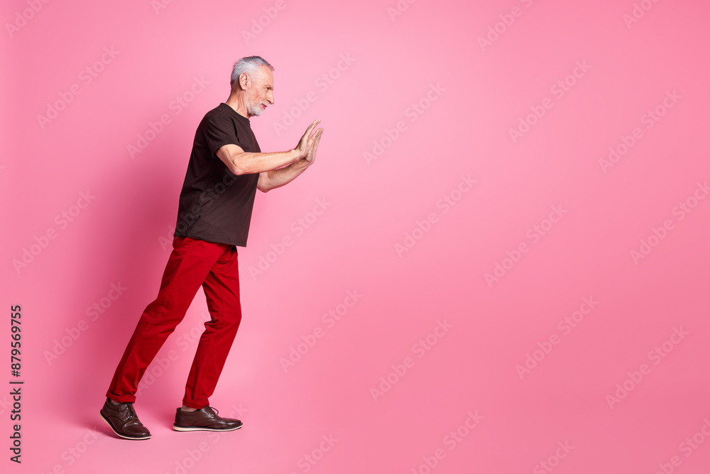Full size photo of serious senior man dressed brown t-shirt red trousers pushing object empty space isolated on pink color background Full size photo of serious senior man dressed brown t-shirt red trousers pushing object empty space isolated on pink color background