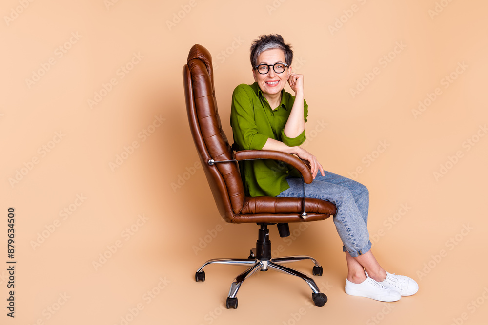 Full size photo of pretty senior female director sit armchair wear trendy green outfit isolated on beige color background Full size photo of pretty senior female director sit armchair wear trendy green outfit isolated on beige color background